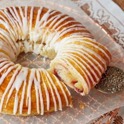 A gorgeous Danish Kringle is golden brown on top gleaming with zig zags of creamy icing. A slice is served on a white dessert plate, showing the swirly and soft inside filled with sweet-tart jam.