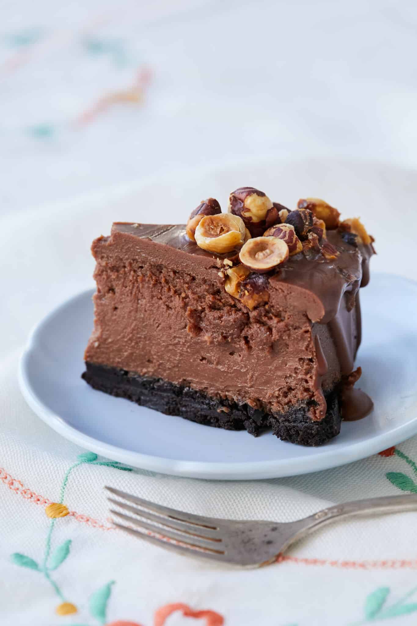 A close-up shot at a slice of the Heavenly Nutella Cheesecake, shows the rich cookie crust, creamy chocolate cream cheese filling and silky smooth ganache coating. It's topped with crunchy toasted hazelnuts for extra flavor and texture. 