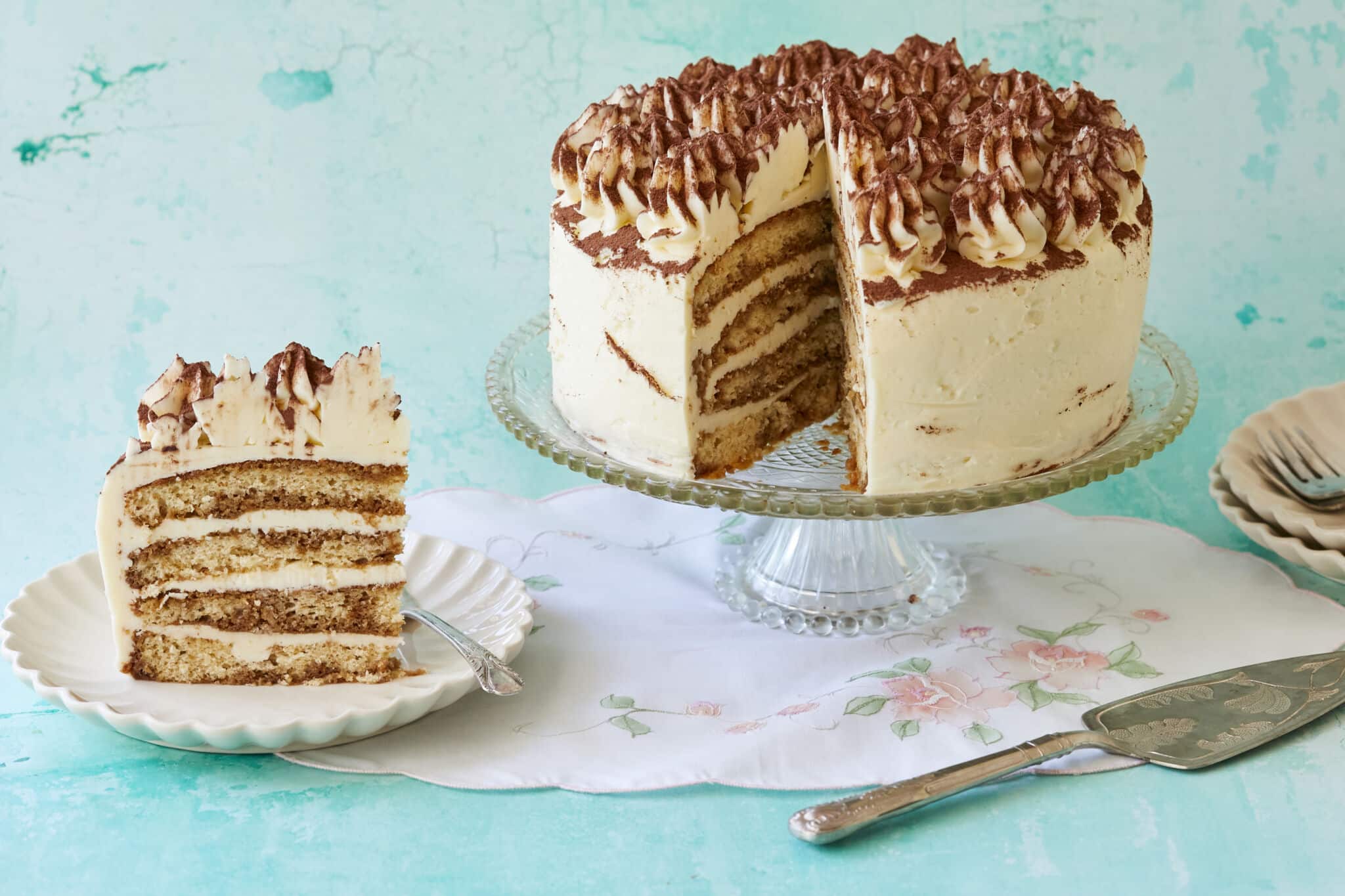 A Stunning Tiramisu Cake is being displayed on a glass cake stand with one generous slice has been cut off. The inside shows its layers of coffee-soaked sponge cake filled with creamy mascarpone. This layered tiramisu cake is topped with rosettes and dusted cocoa which makes it visually appealing.