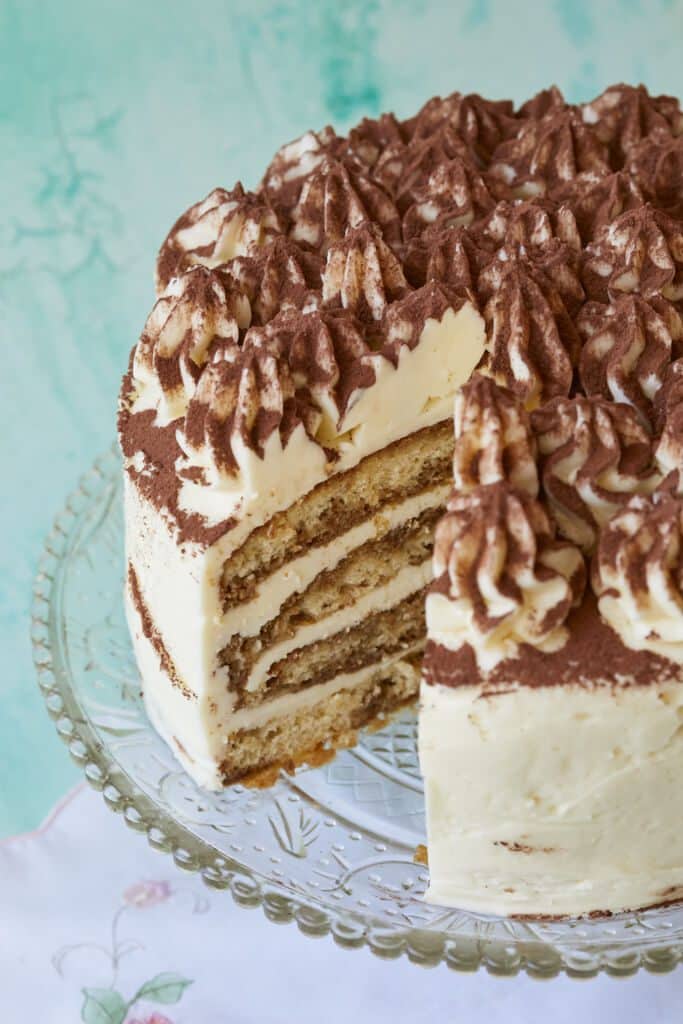 A Stunning Tiramisu Cake is served on a glass cake stand with one generous slice cut off. The inside shows its layers of coffee-soaked sponge cake filled with creamy mascarpone. This layered tiramisu cake is topped with rosettes and dusted cocoa which makes it visually appealing.