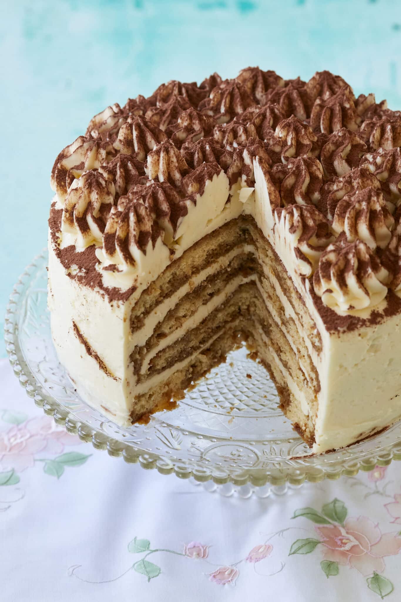 A Stunning Tiramisu Cake is being displayed on a glass cake stand with one generous slice has been cut off. The inside shows its layers of coffee-soaked sponge cake filled with creamy mascarpone. This layered tiramisu cake is topped with rosettes and dusted cocoa which makes it visually appealing. 