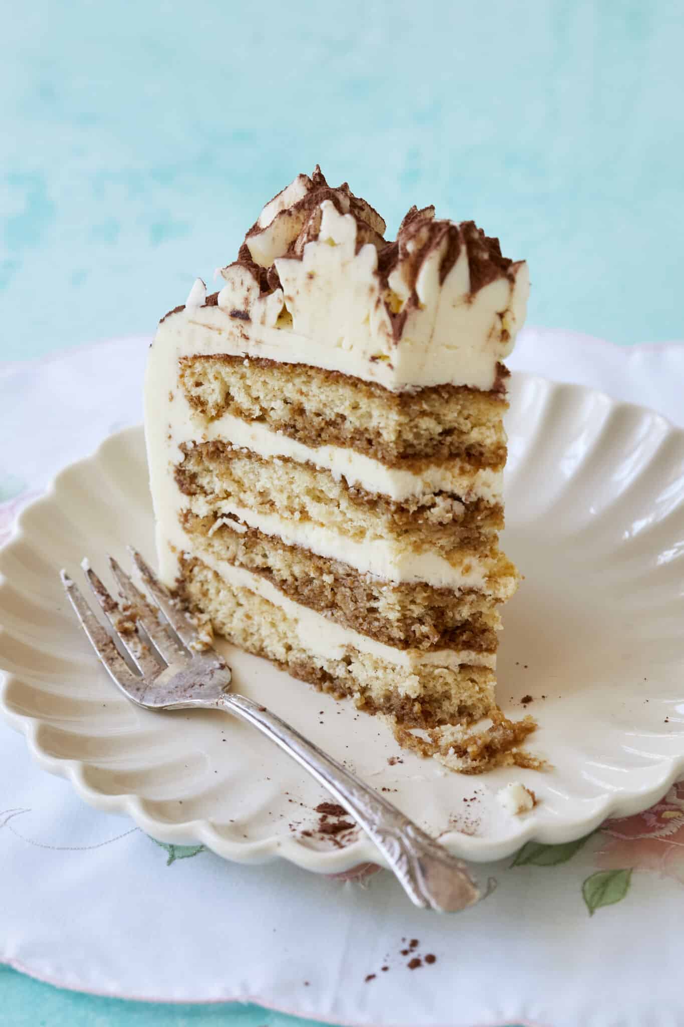 The close-up shot at a slice of the Stunning Tiramisu Cake served on a dessert plate shows its layers of coffee-soaked sponge cake filled with creamy mascarpone. This layered tiramisu cake is topped with rosettes and dusted cocoa which makes it visually appealing. 