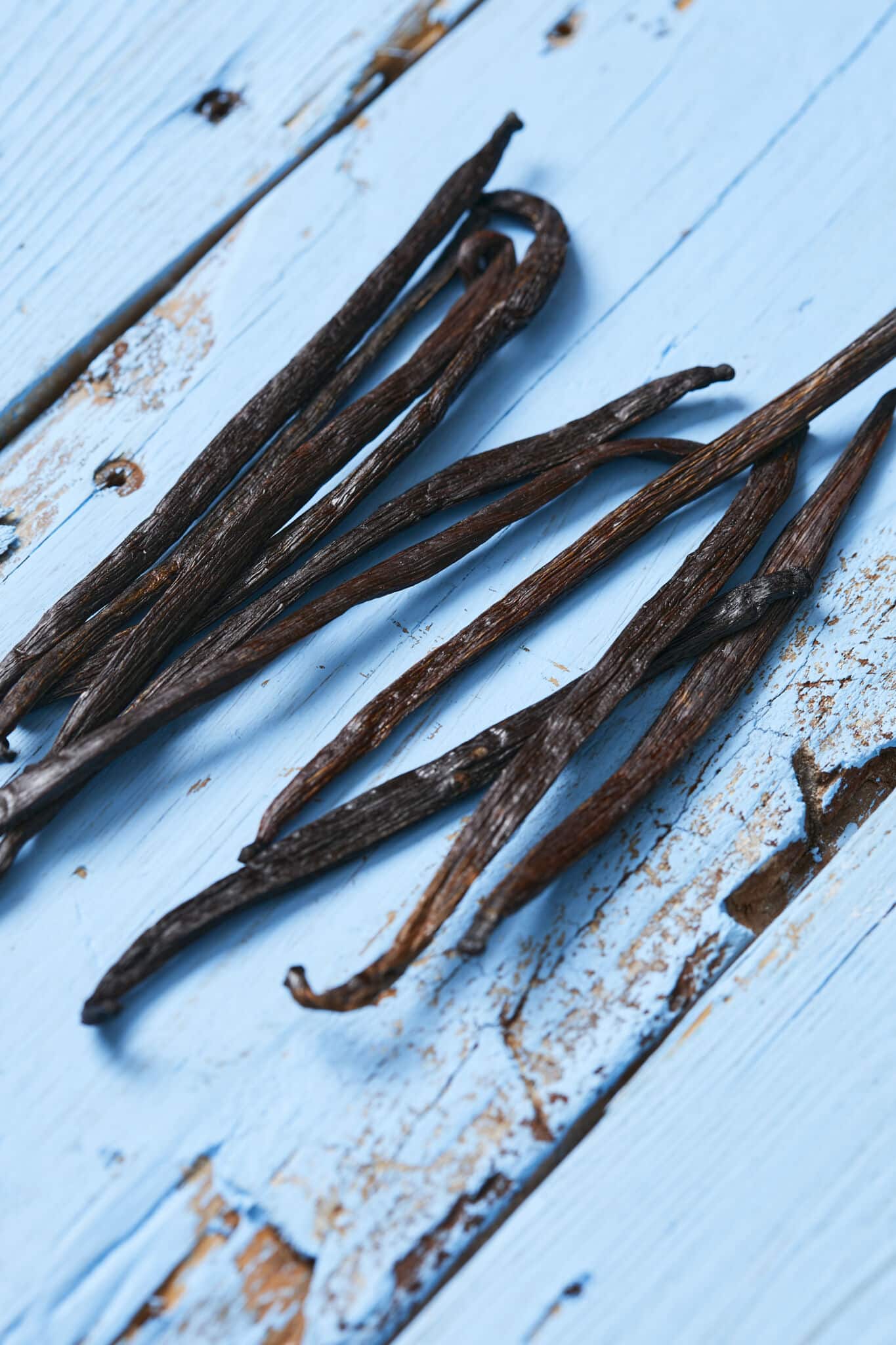 Long and slender vanilla bean pods are placed on a blue wooden board, resembling thin dark brown or blackish-brown cylinders. They are about 4 to 8 inches (10 to 20 centimeters) in length. with a wrinkled, leathery surface texture.