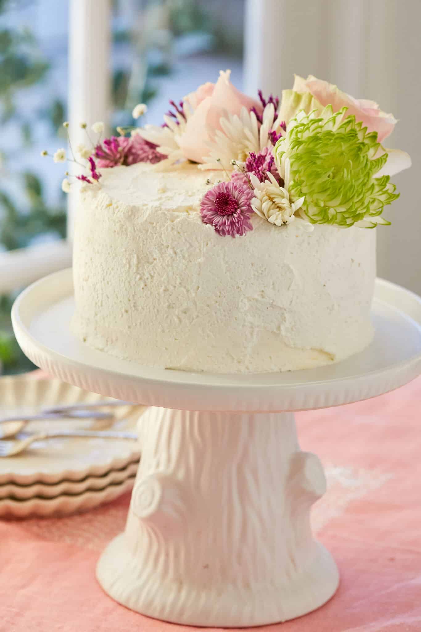 The Vertical Lemon Cake is a visually stunning, covered with white mascarpone whipped cream and topped with fresh colorful flowers . It's being displayed on a white tree-stump cake stand.