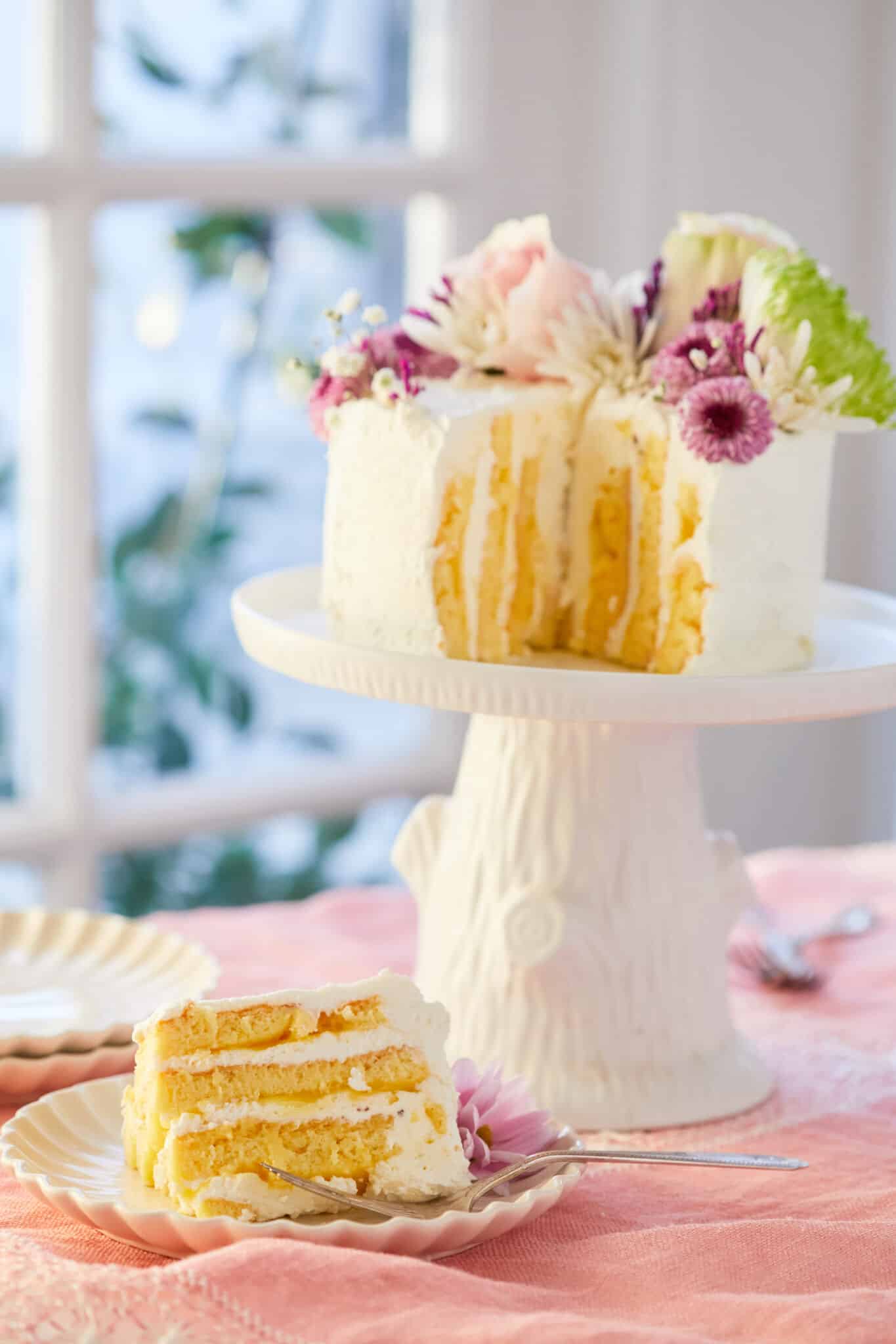 A stunning Vertical Lemon Cake is being displayed on a white tree-stump cake stand, covered with white mascarpone whipped cream and topped with colorful fresh flowers. One slice is cut off and served on a dessert plate. The slice shows the layers of soft cake covered with luscious lemon curd and mascarpone whipped cream on the inside and generously frosted with additional mascarpone whipped cream on the outside. 