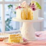 A stunning Vertical Lemon Cake is being displayed on a white tree-stump cake stand, covered with white mascarpone whipped cream and topped with fresh flowers. One slice is cut off and served on a dessert plate. The slice shows the layers of soft cake covered with luscious lemon curd and mascarpone whipped cream on the inside and generously frosted with additional mascarpone whipped cream on the outside.