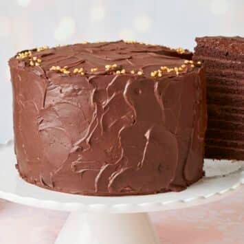 A generous slice of the 24-Layer Chocolate Cake is being lifted from the platter. A close-up shot at the slice shows its moist and soft crumb and silky smooth ganache on the outside and decadent chocolate pastry cream between the layers.