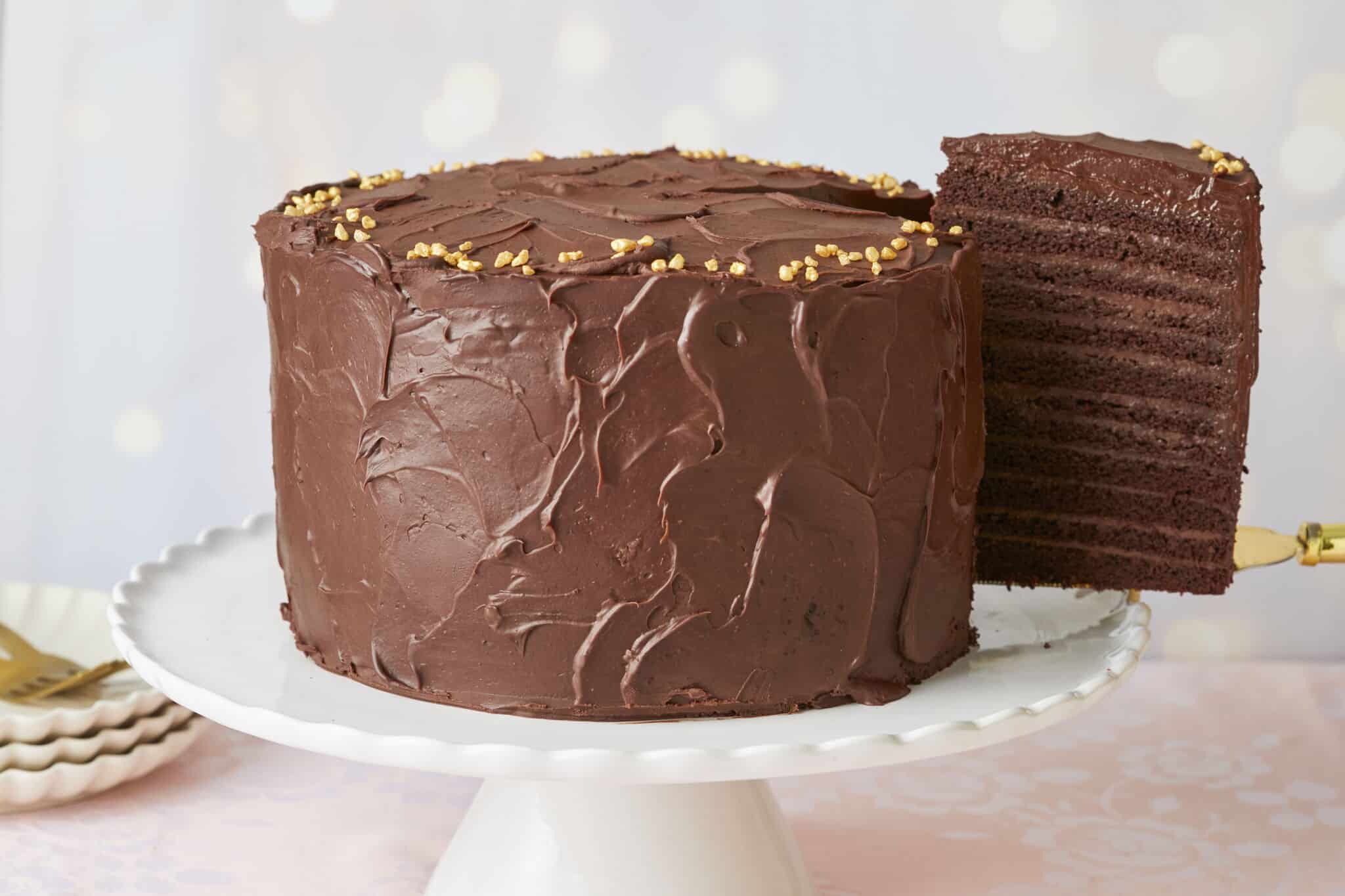 A generous slice of the 24-Layer Chocolate Cake is being lifted from the platter. A close-up shot at the slice shows its moist and soft crumb and silky smooth ganache on the outside and decadent chocolate pastry cream between the layers.