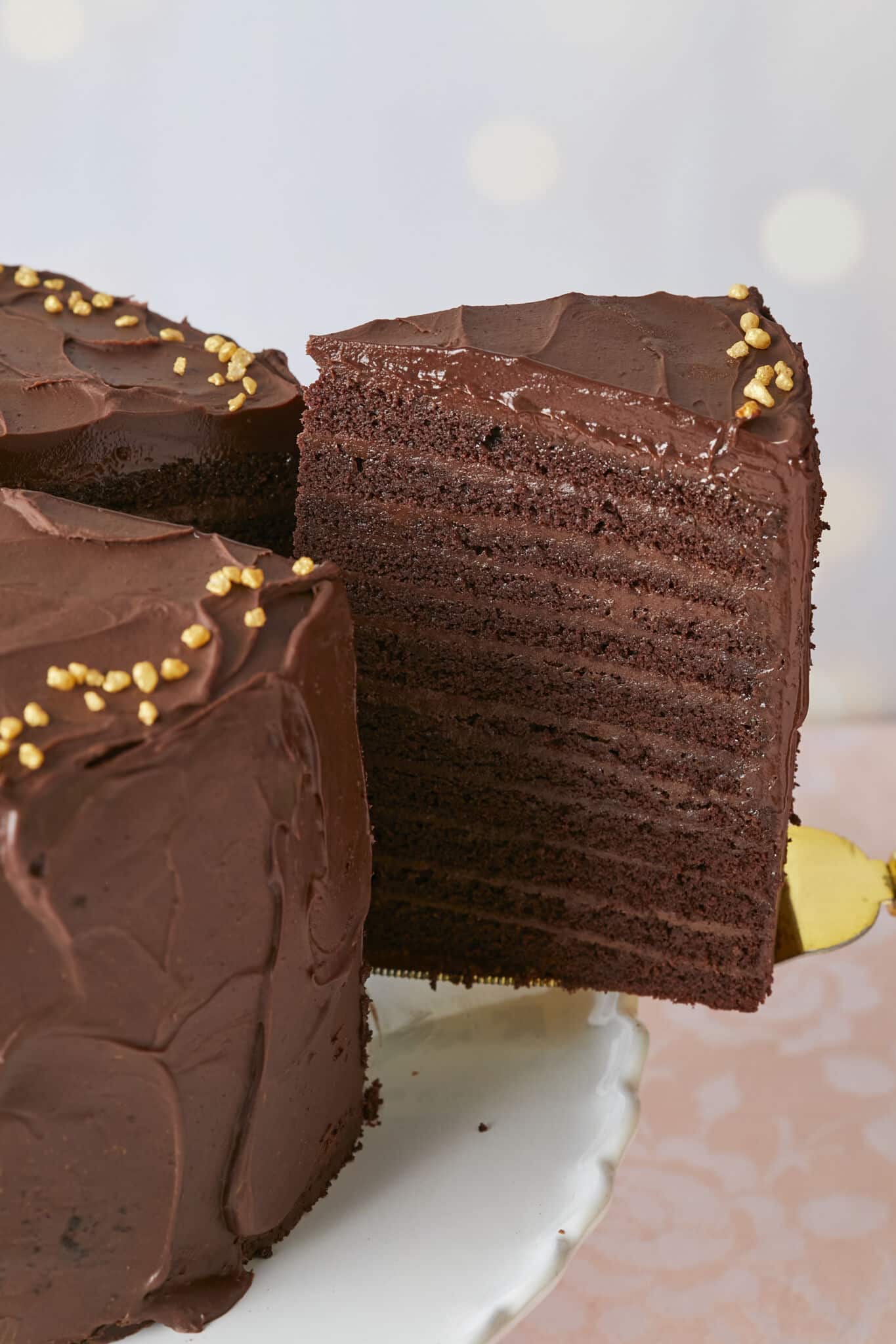 A generous slice of the 24-Layer Chocolate Cake is being lifted from the platter. A close-up shot at the slice shows its moist and soft crumb and silky smooth ganache on the outside and decadent chocolate pastry cream between the layers. 