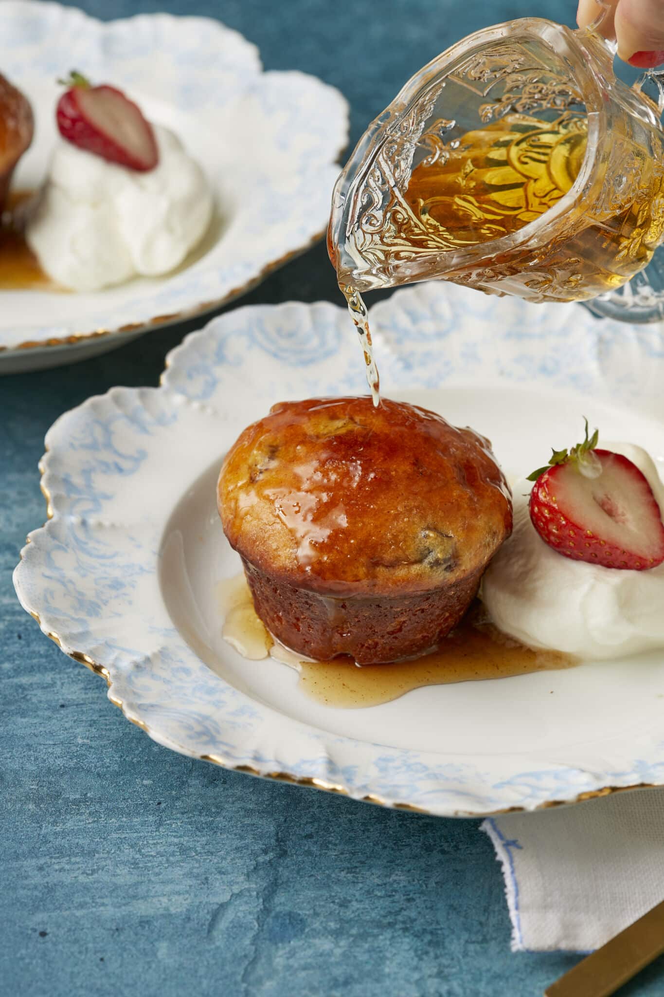 Two soft, delicate Baba au Rhum (Classic Rum Baba) cakes are served with fresh strawberry and whipped cream. Syrup is being drizzled from top to bottom.