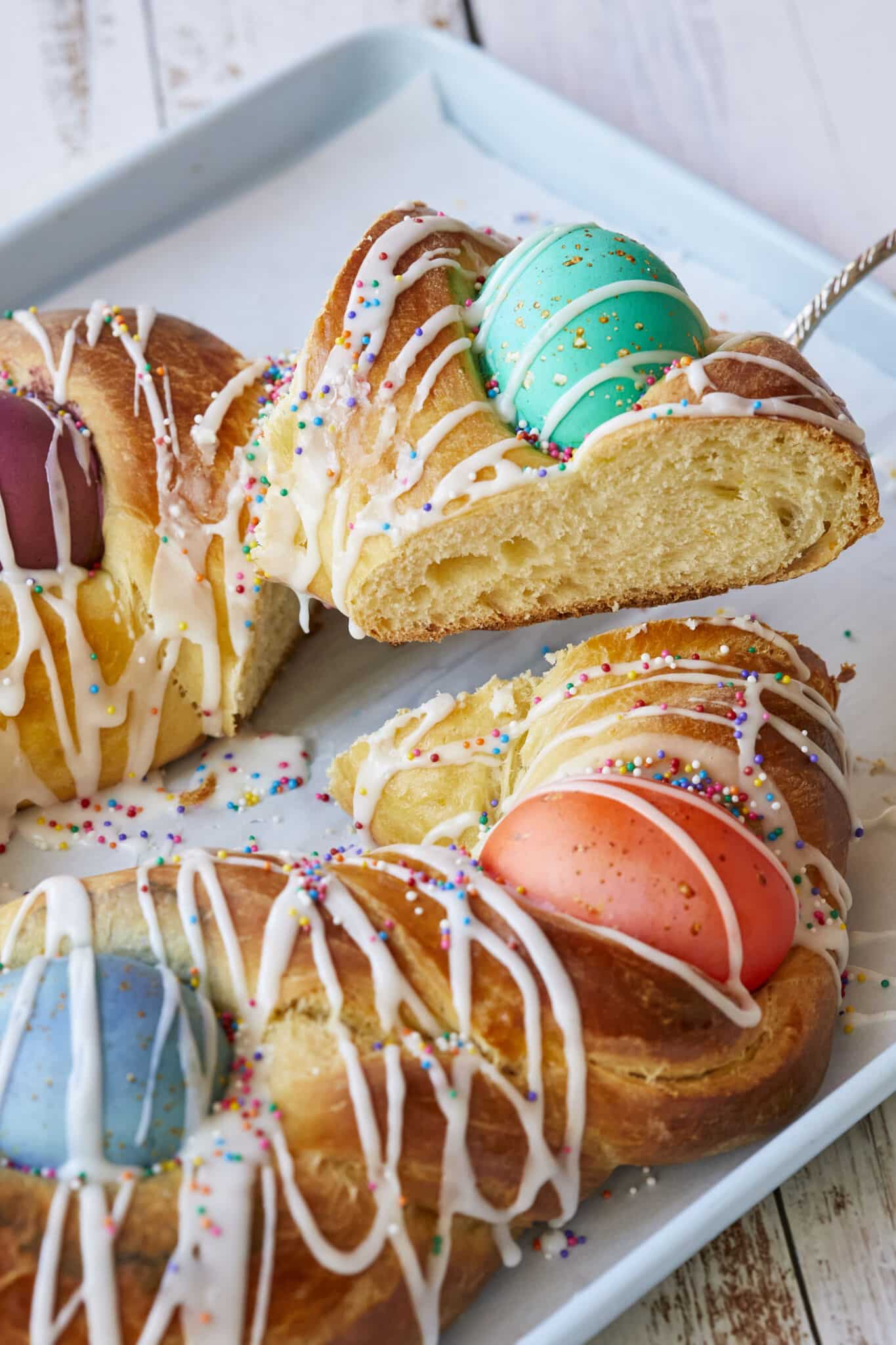 A close-up shot at the Traditional Italian Easter Bread (Pane di Pasqua) . A slice is cut and being lifted from the pan. The bread is golden, light and fluffy, and decorated with colorful Easter eggs, drizzled with icing and topped with colorful sprinkles. 