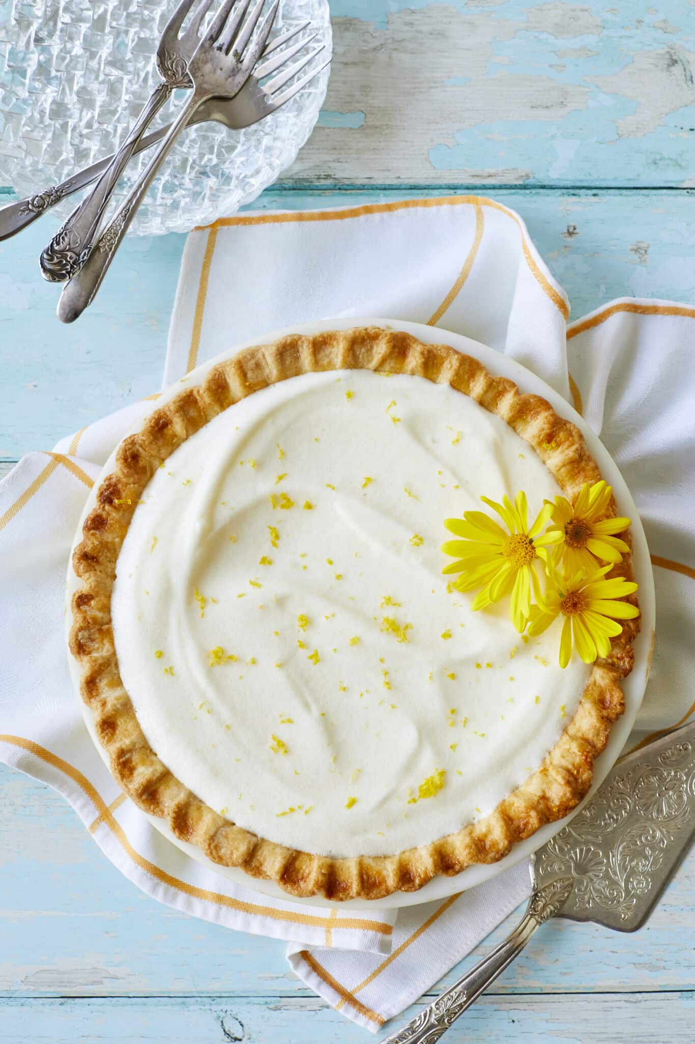An over-head shot shows that the Luscious Creamy Lemon Chiffon Pie has a golden curst with a white, silky filling, topped with refreshing lemon zest and two flowers. 