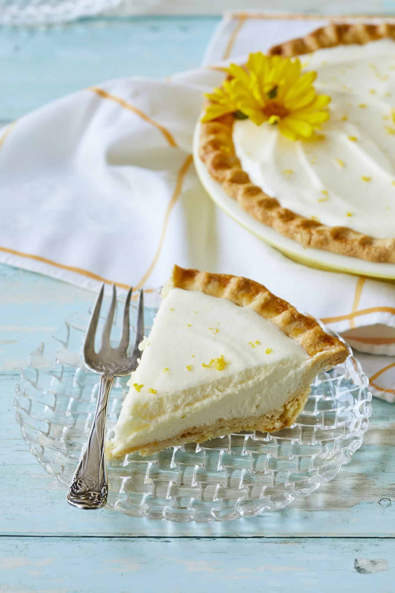 Luscious Creamy Lemon Chiffon Pie has a golden curst with a white, silky filling, topped with refreshing lemon zest and two flowers. A slice is served on a glass dessert plate, showing the flaky crust and smooth filling.