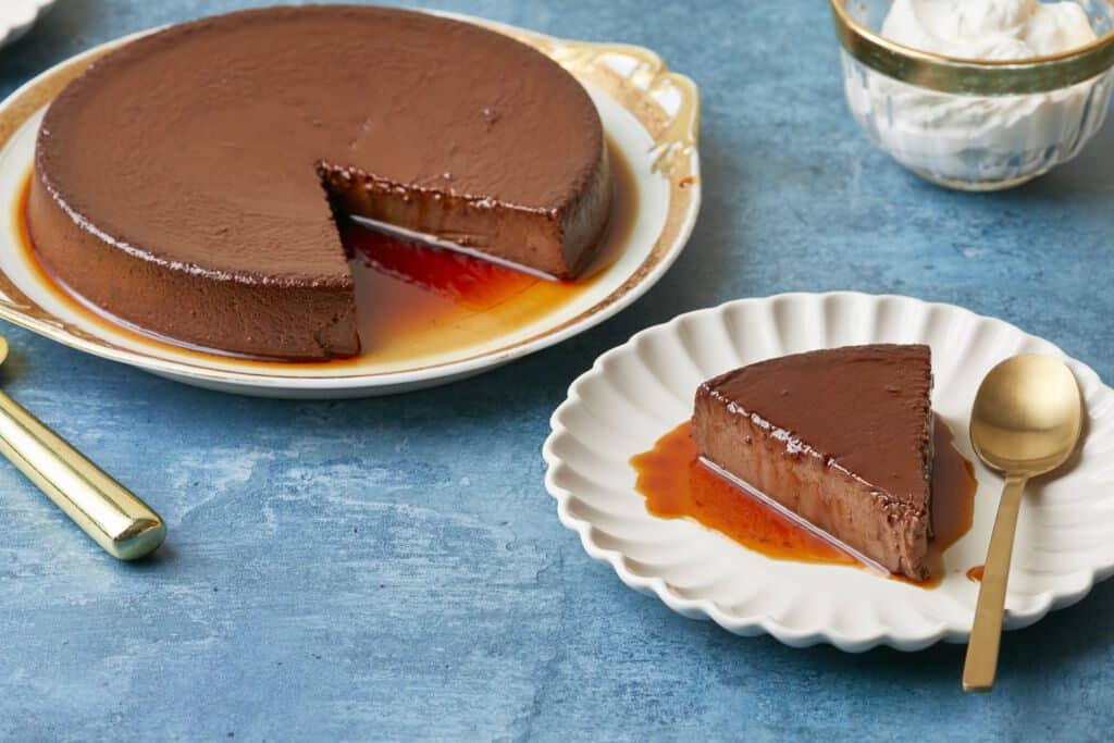 Mexican Chocolate Flan is served on a golden rim platter and a slice has been cut and served on a dessert plate. The flan is silky smooth in deep chocolate color and covered with a thin layer of caramel.
