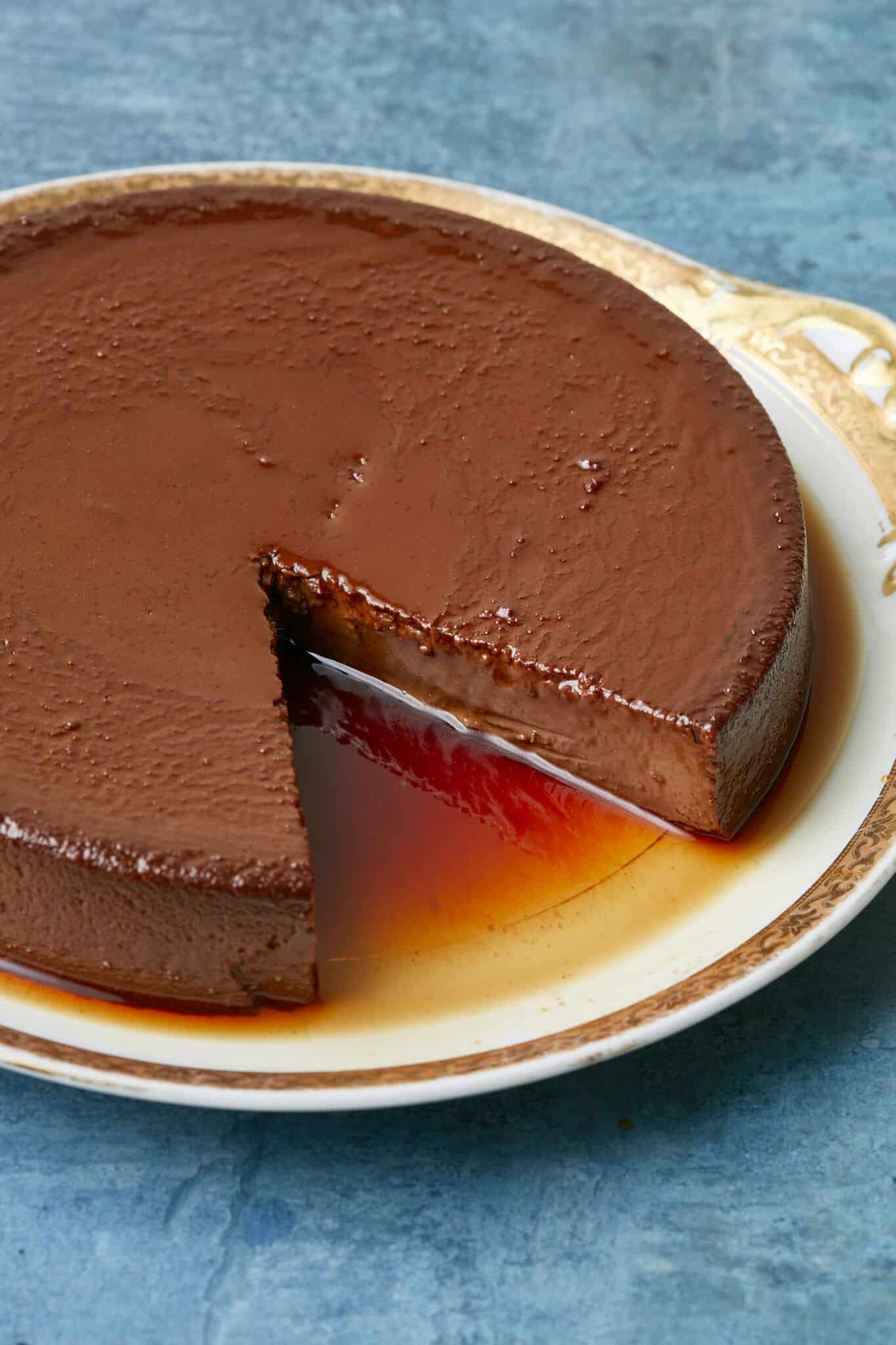 A slice has been cur from the Mexican Chocolate Flan that's served on a golden rim platter. The close-up shot shows that it is silky smooth in deep chocolate color and covered with a thin layer of caramel. 