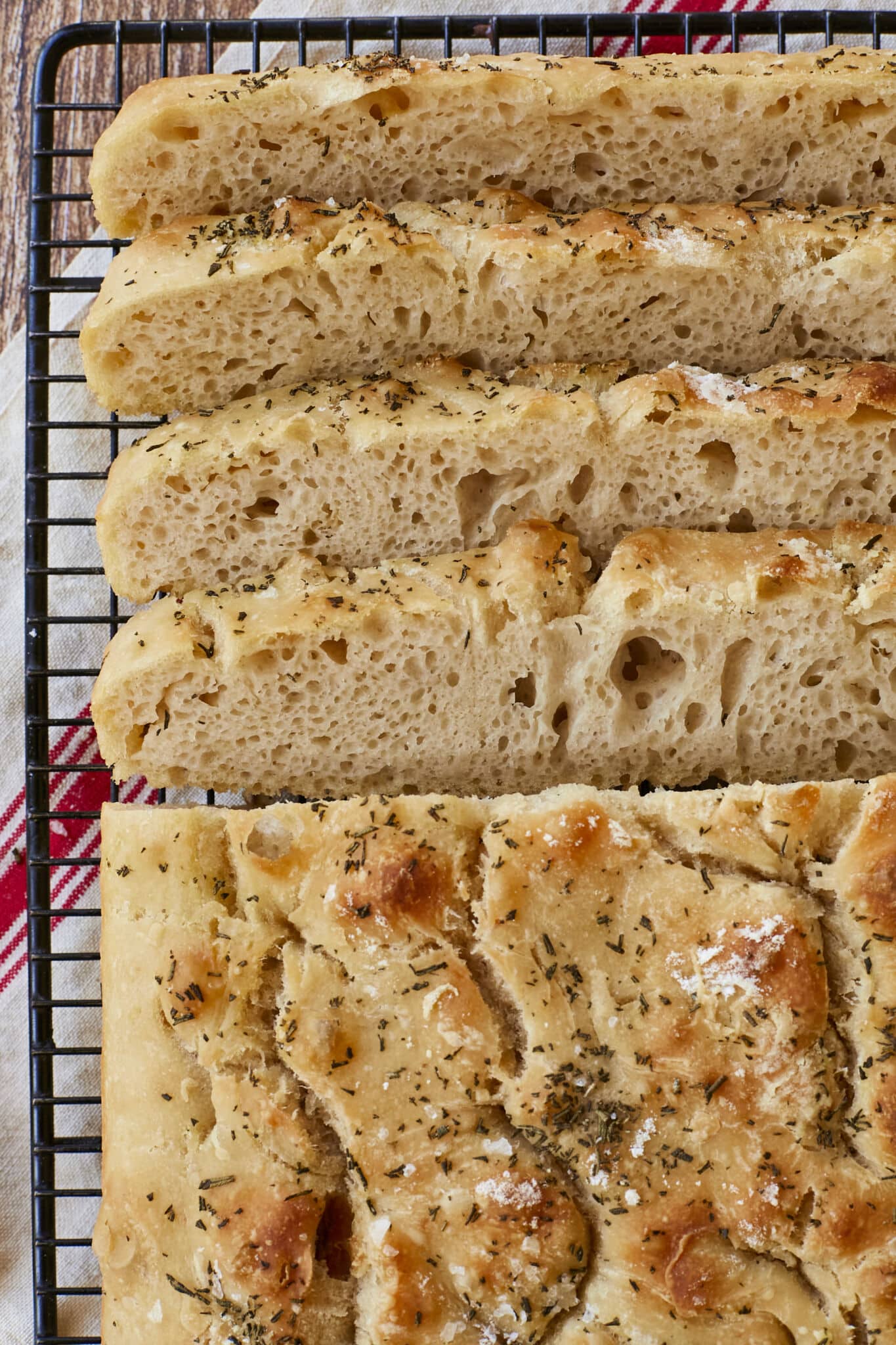 No-knead focaccia is sliced and cooling on the black wire rack. The close-up shot shows it bubbly crumb and golden, crispy exterior loaded with flaky sea salt, Roasted Garlic and Rosemary. 