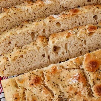 No-knead focaccia is sliced and cooling on the black wire rack. The close-up shot shows it bubbly crumb and golden, crispy exterior loaded with flaky sea salt, Roasted Garlic and Rosemary.