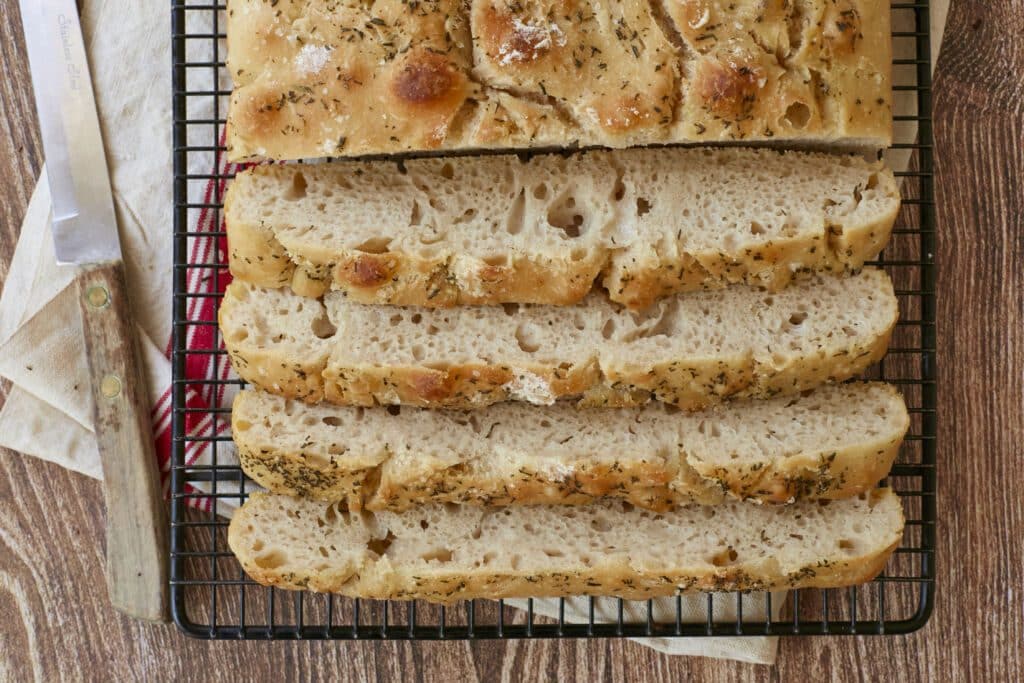 No-knead focaccia is sliced and cooling on the black wire rack. The close-up shot shows it bubbly crumb and golden, crispy exterior loaded with flaky sea salt, Roasted Garlic and Rosemary.