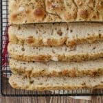 No-knead focaccia is sliced and cooling on the black wire rack. The close-up shot shows it bubbly crumb and golden, crispy exterior loaded with flaky sea salt, Roasted Garlic and Rosemary.