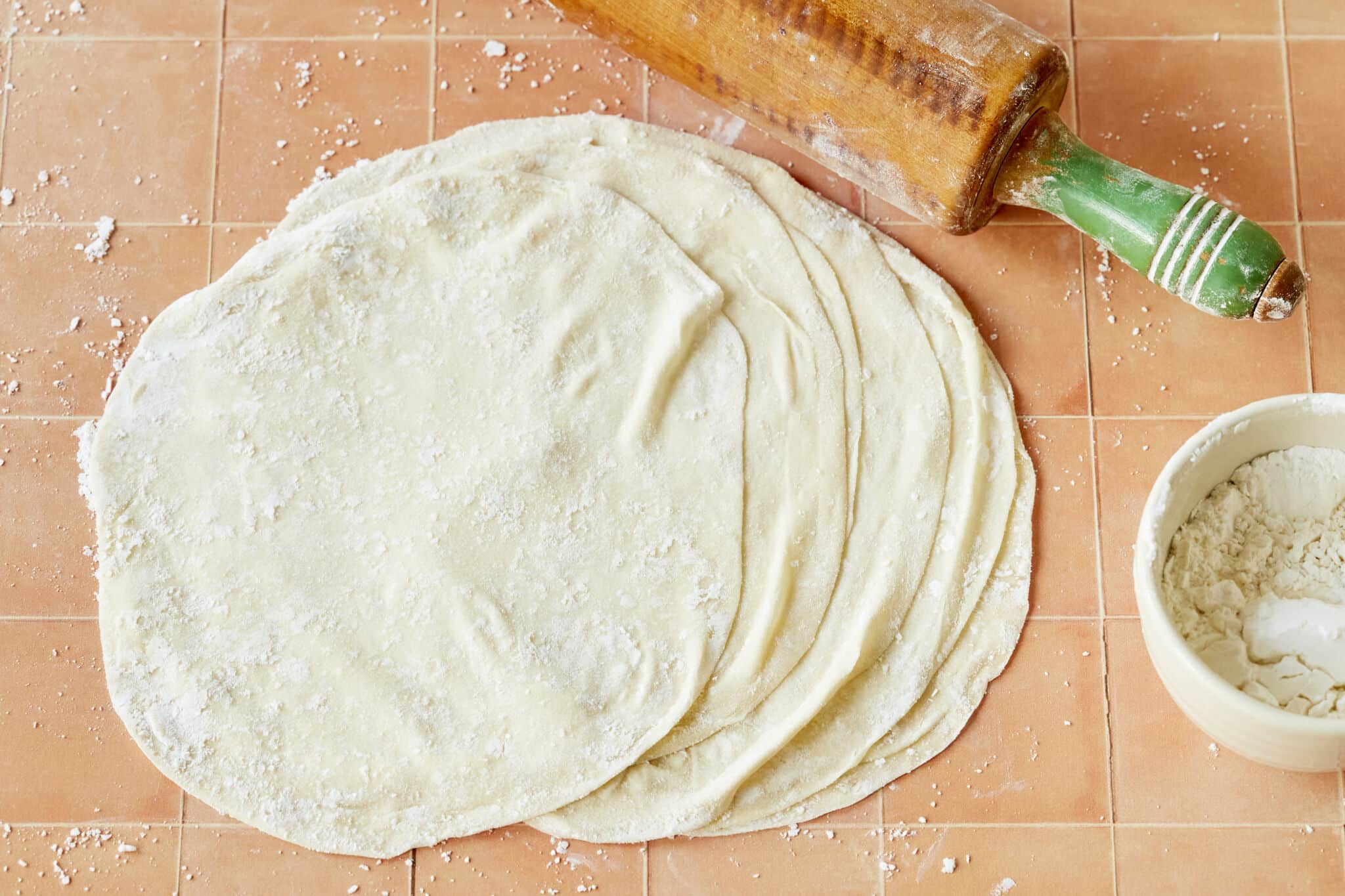 A stack of delicate, paper-thin Homemade Phyllo Dough (Fill Pastry) is dusted with flour. A bowl of extra flour and a rolling pin are on the side.