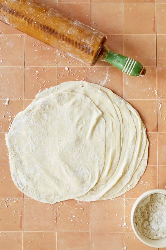 A stack of delicate, paper-thin Homemade Phyllo Dough (Fill Pastry) is dusted with flour. A bowl of extra flour and a rolling pin are on the side.