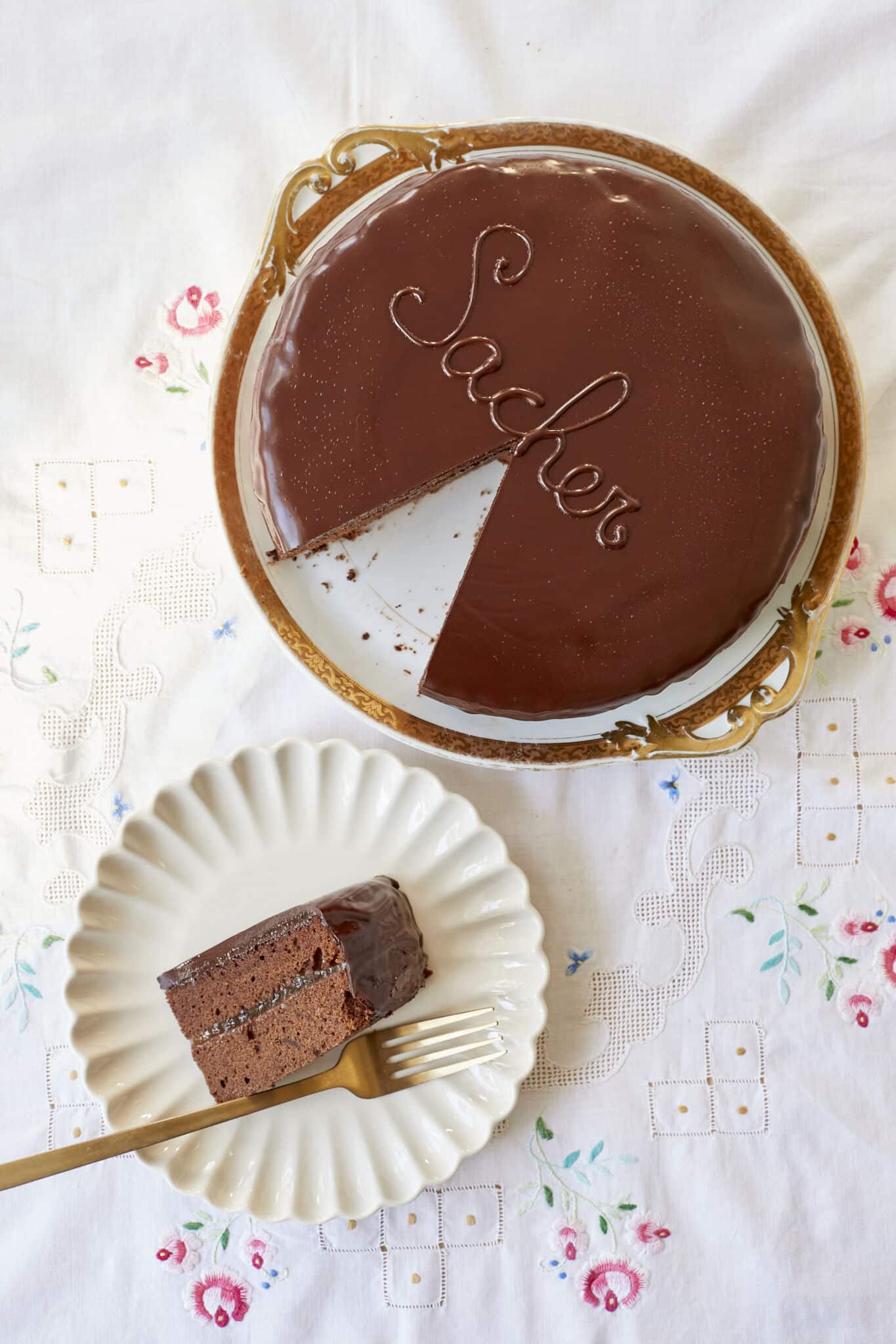 Classic Austrian Sacher Torte is placed on a round golden rim platter. A slice is cut and served on a white dessert plate. It shows the moist, fine crumb of the chocolate sponge cake and the silky smooth, shinny glaze. 
