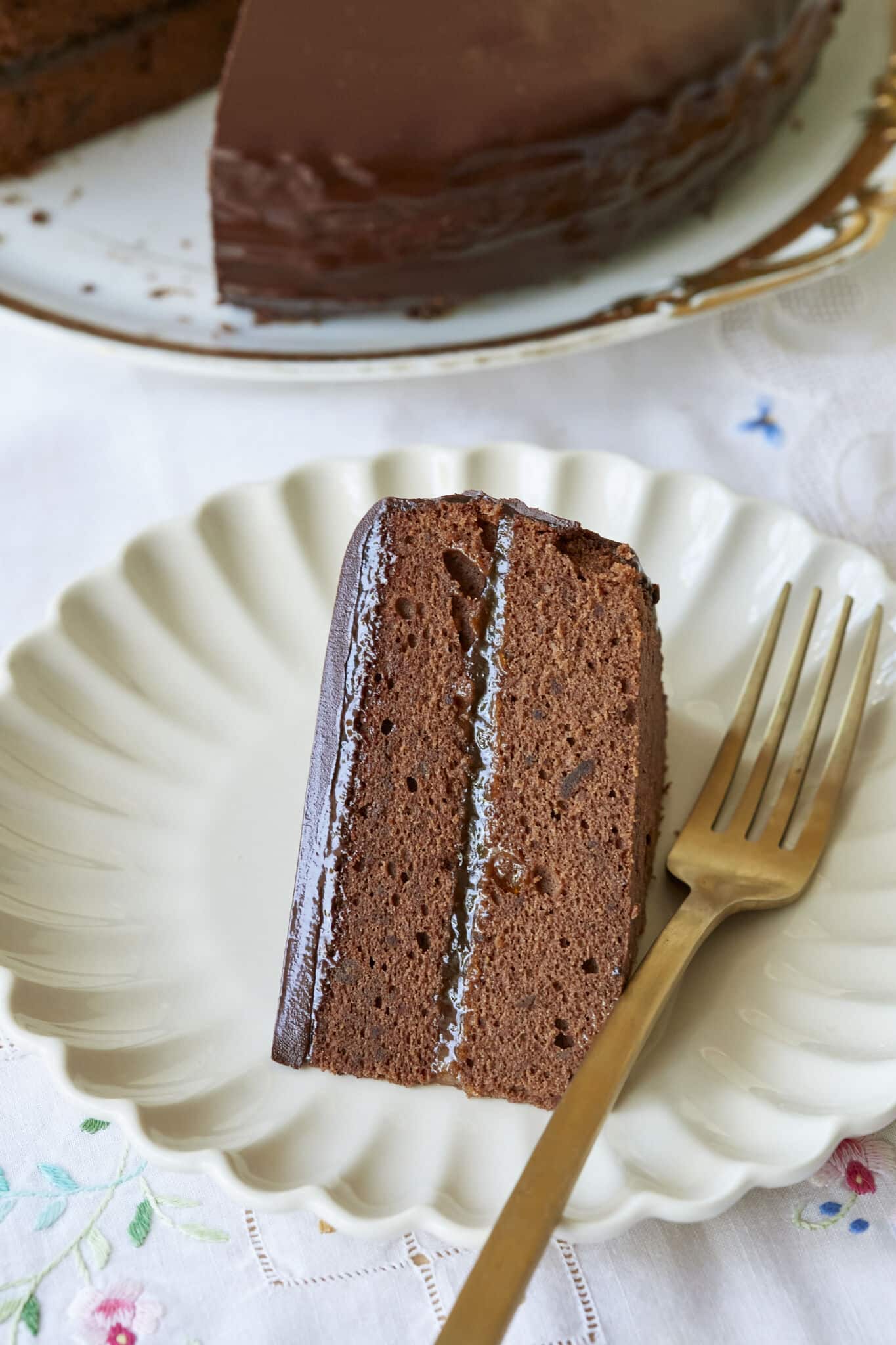 Classic Austrian Sacher Torte is placed on a round golden rim platter. A case-up shot at the slice on a white dessert plate shows the moist, fine crumb of the chocolate sponge cake and the silky smooth, shinny glaze. 