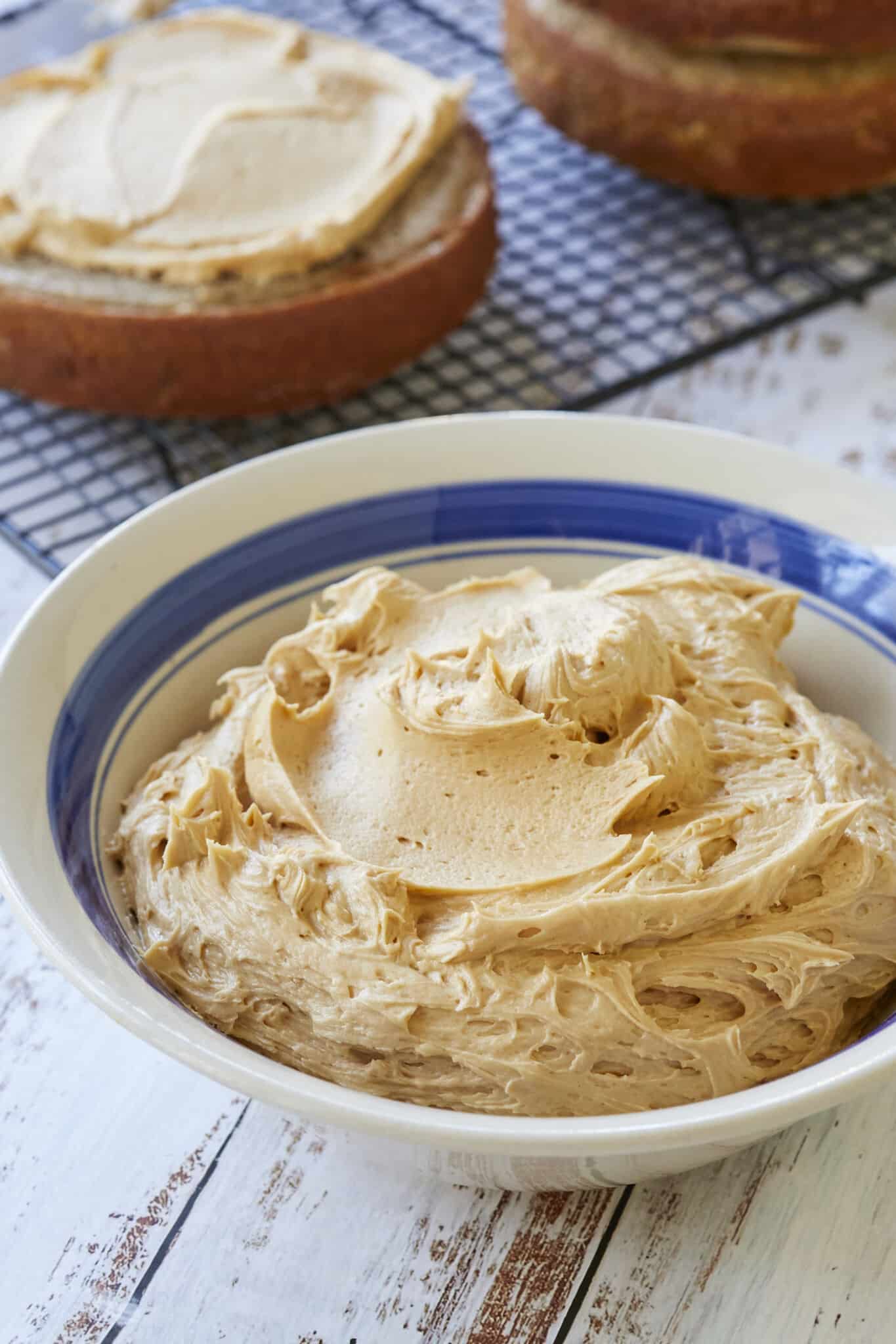 A close-up shot at a big bowl of smooth Salted Caramel Buttercream Frosting shows its creamy texture. Two round cake layers are on the cooling rack. One has been frosted. 