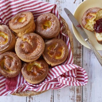 A basket of golden, light and fluffy Sourdough Popovers. One popover is sliced open and served on a dessert plate with jam.