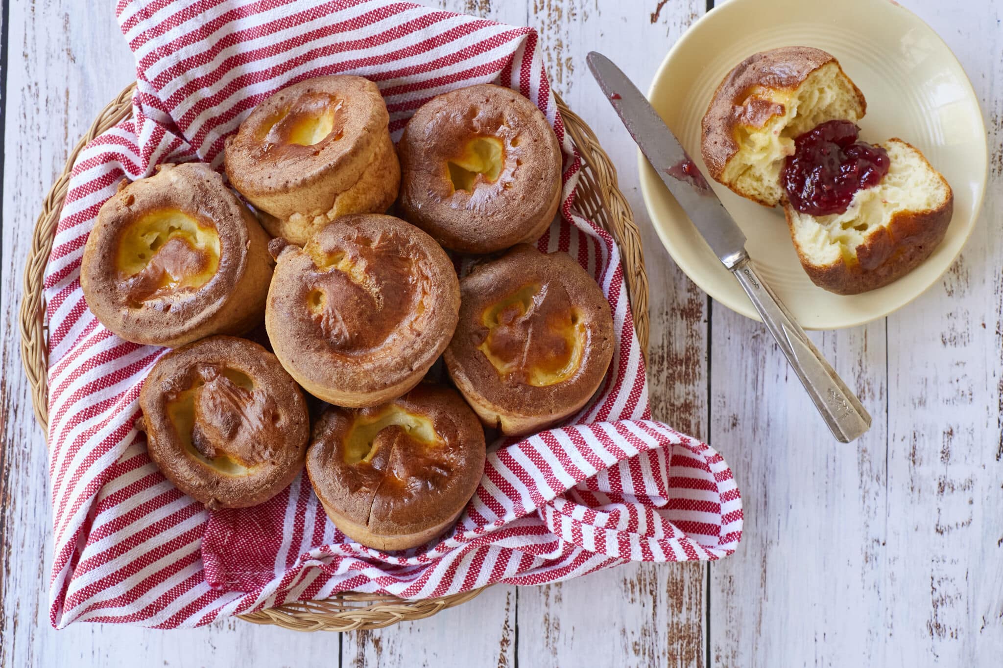 A basket of golden, light and fluffy Sourdough Popovers. One popover is sliced open and served on a dessert plate with jam.