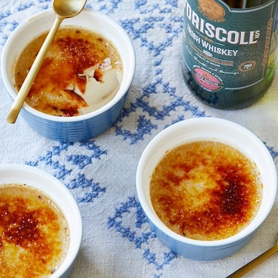 An overhead shot of 3 ramekins of Irish Whiskey Crème Brûlée shows the caramelized crunch top and silky smooth custard interior. A bottle of Irish Whiskey is on the side.