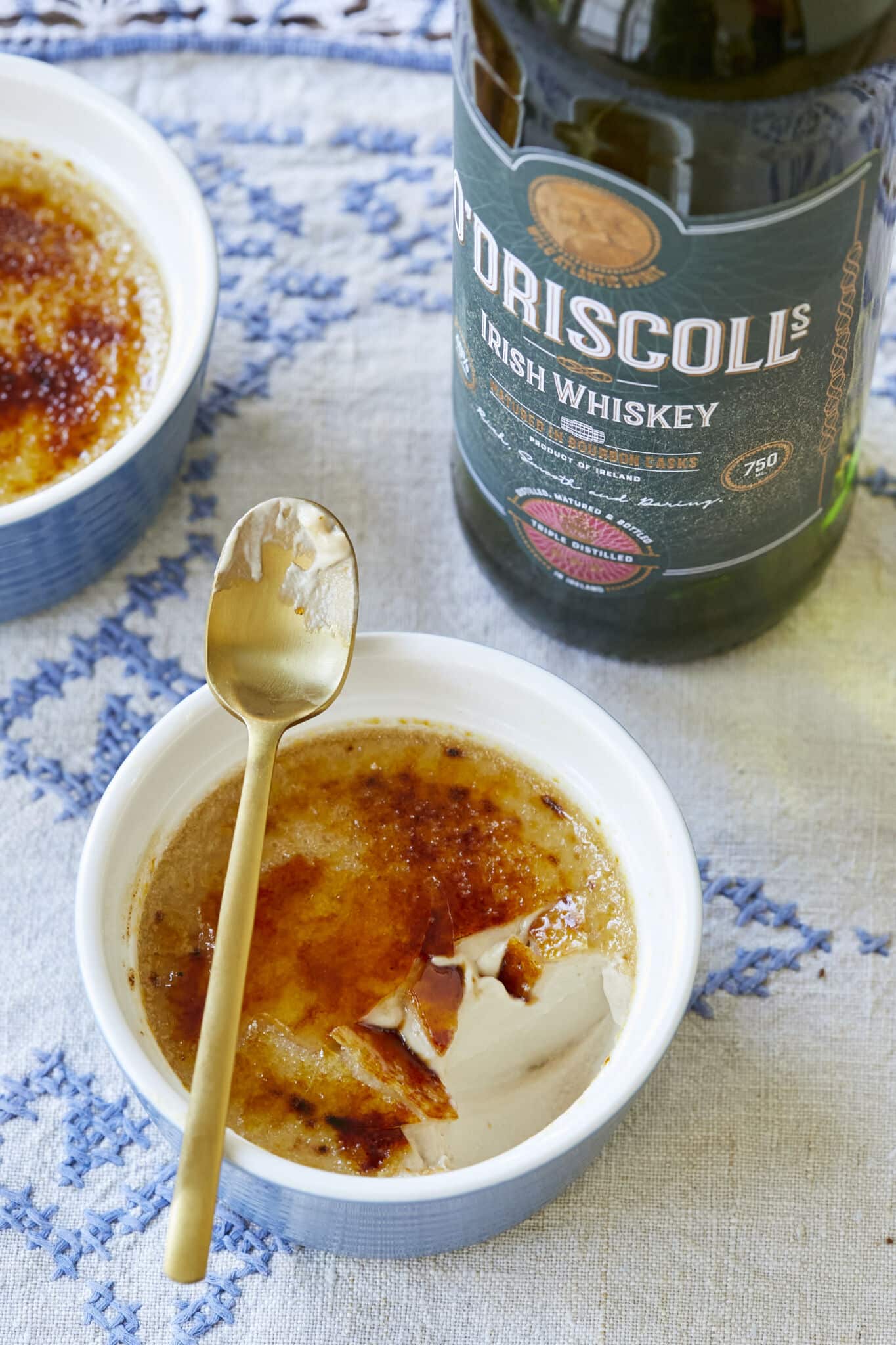 A close-up shot of Irish Whiskey Crème Brûlée shows the caramelized crunch top and silky smooth custard interior. A bottle of Irish Whiskey is on the side. 