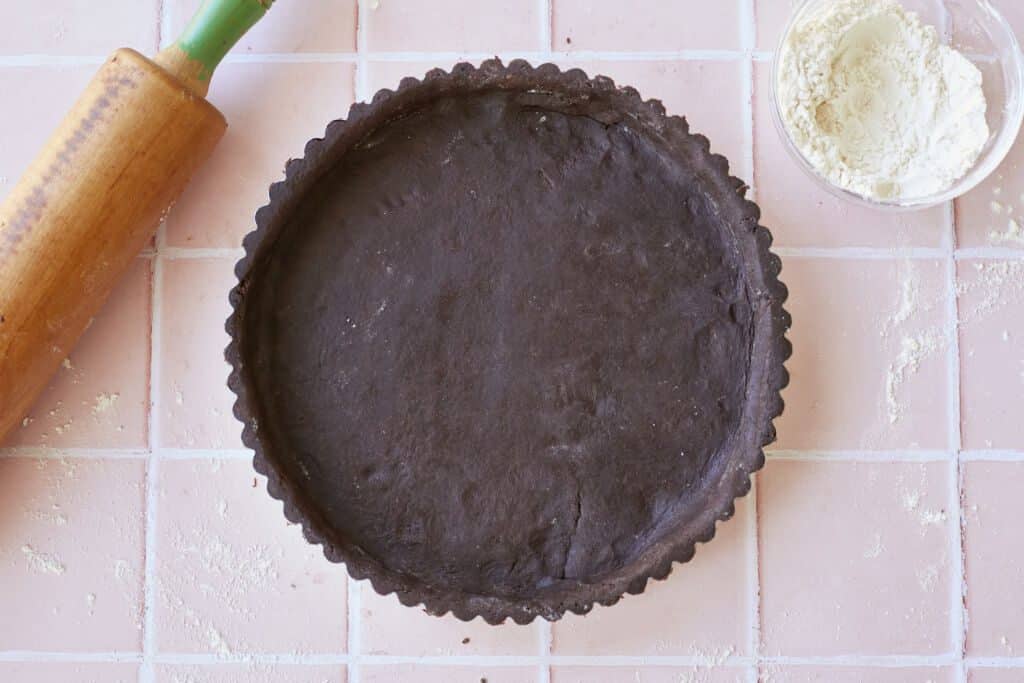 Chocolate Pastry is perfectly prepared in the fluted-edge pie pan. A wooden rolling pin is in the top left corner of the image and a small bowl of extra flour is in the top right corner.