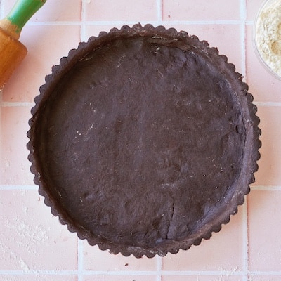 Chocolate Pastry is perfectly prepared in the fluted-edge pie pan. A wooden rolling pin is in the top left corner of the image and a small bowl of extra flour is in the top right corner.