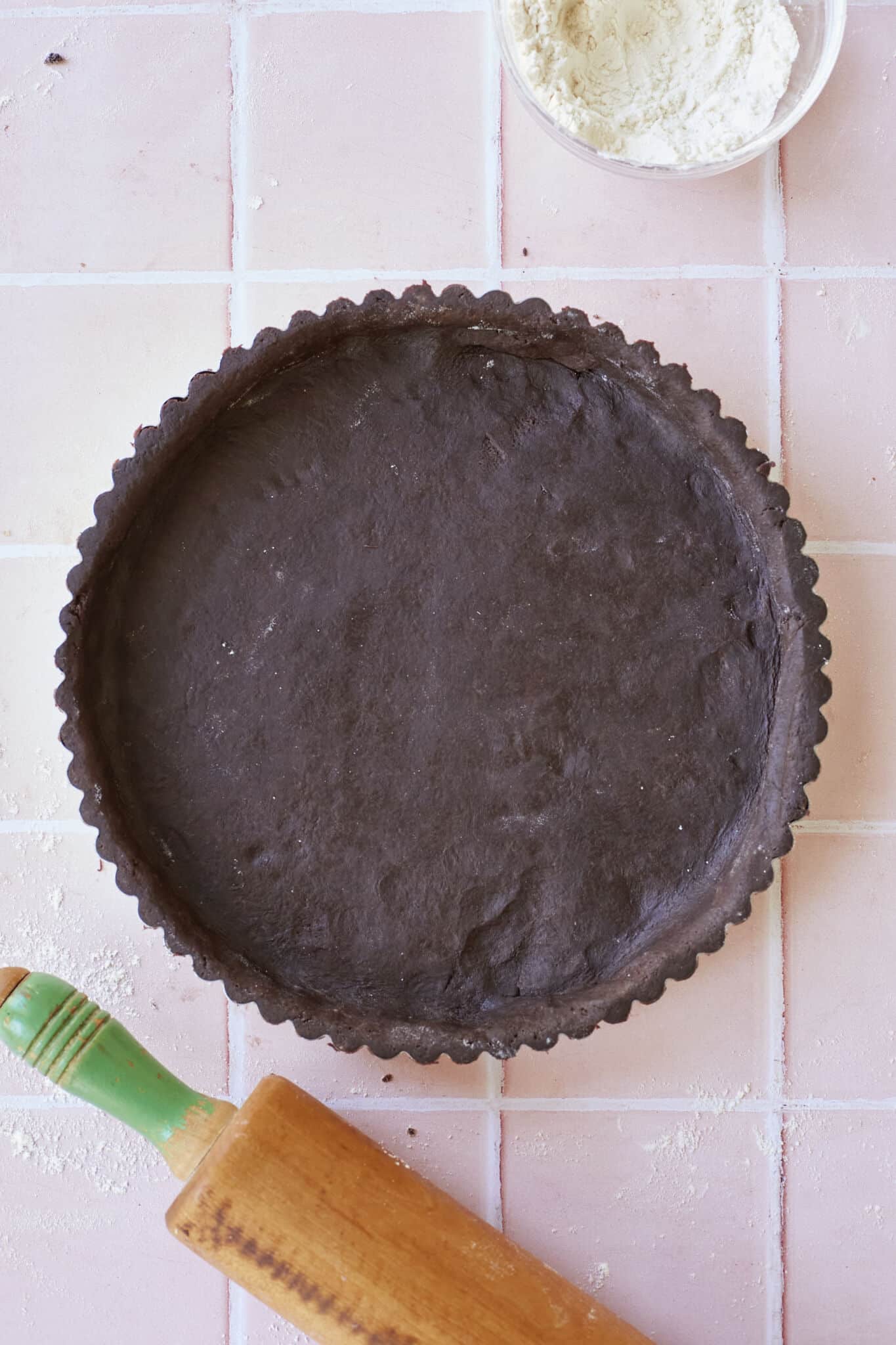 Chocolate Pastry is perfectly prepared in the fluted-edge pie pan. A wooden rolling pin is in the left bottom corner of the image and a small bowl of extra flour is in the top right corner. 