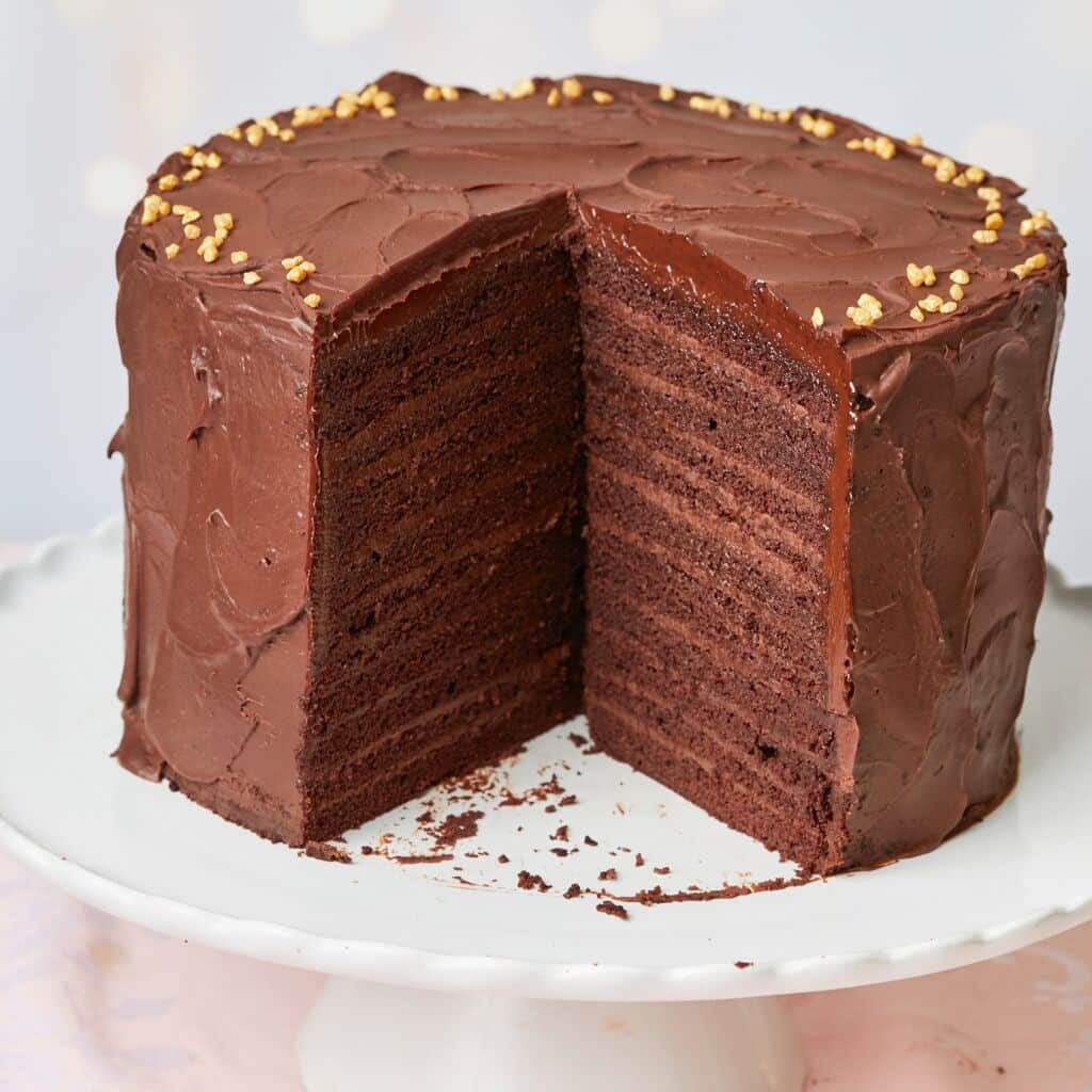 A luxurious 24-Layer Chocolate Cake is served on a cake stand. A generous slice has been cut. The close-up shot at the big cake shows its moist and soft crumb and silky smooth ganache on the outside and decadent chocolate pastry cream between the layers.