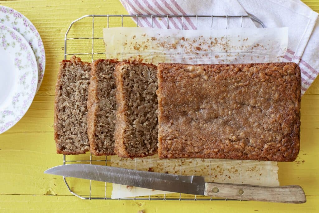 A loaf of golden, moist, soft Almond Banana Bread is sliced and cooling on the wire rack.