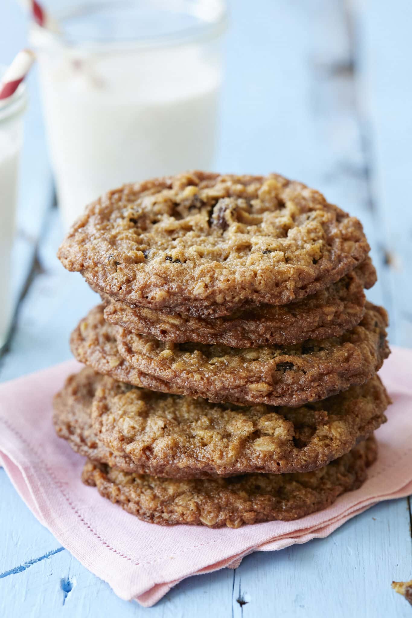 A close-up shot at a stack of Almond Flour Cinnamon Raisin Cookies with glasses of milk in the background. The cookies are golden brown with dark, chewy and crispy edges. 
