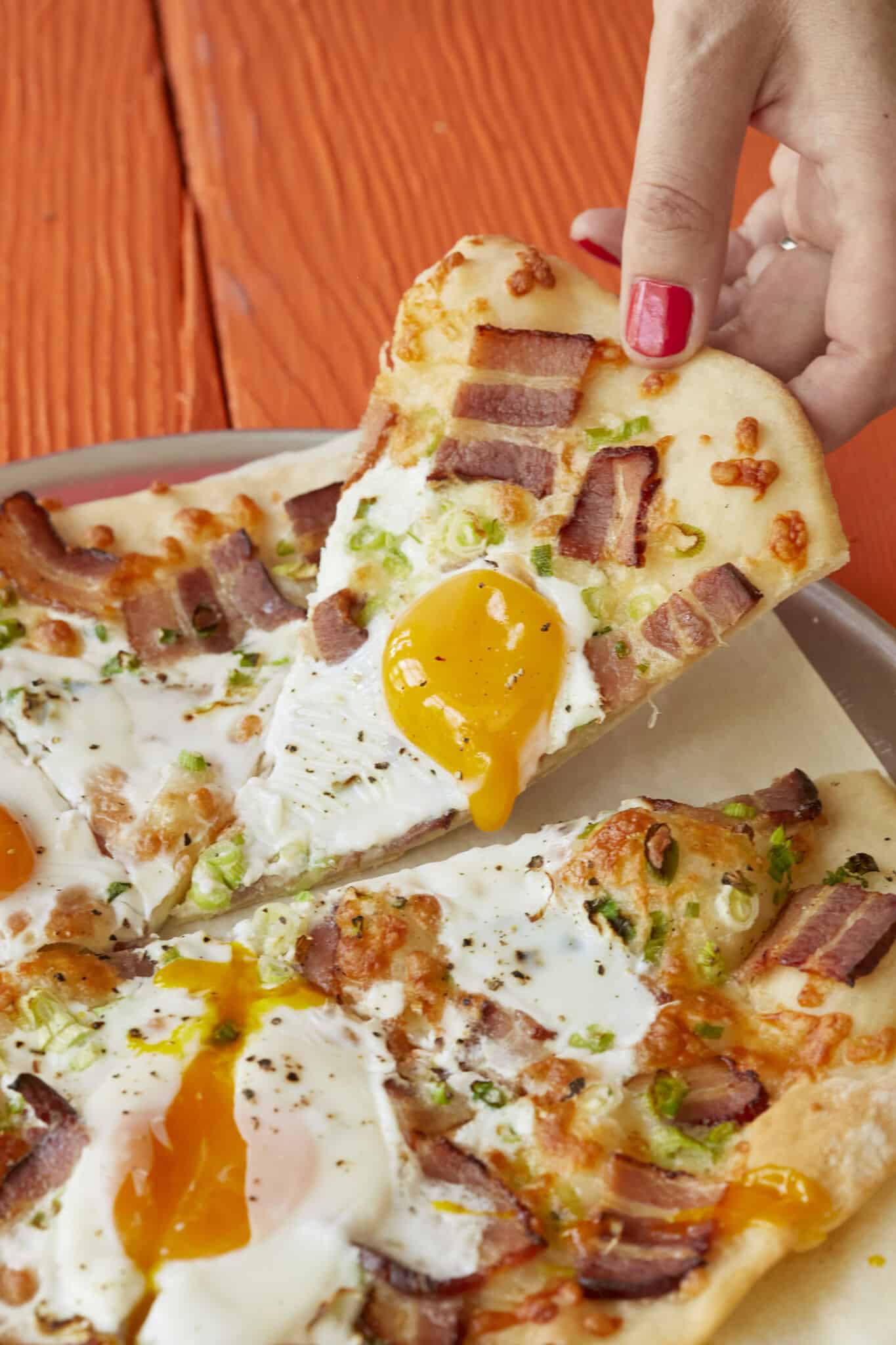 Wholesome breakfast pizza has golden and crispy crust, with smoky bacon, green onions, and sunny-side-up eggs bringing layers of savory flavor. It's sliced and one slice is being lifted from the pan. 