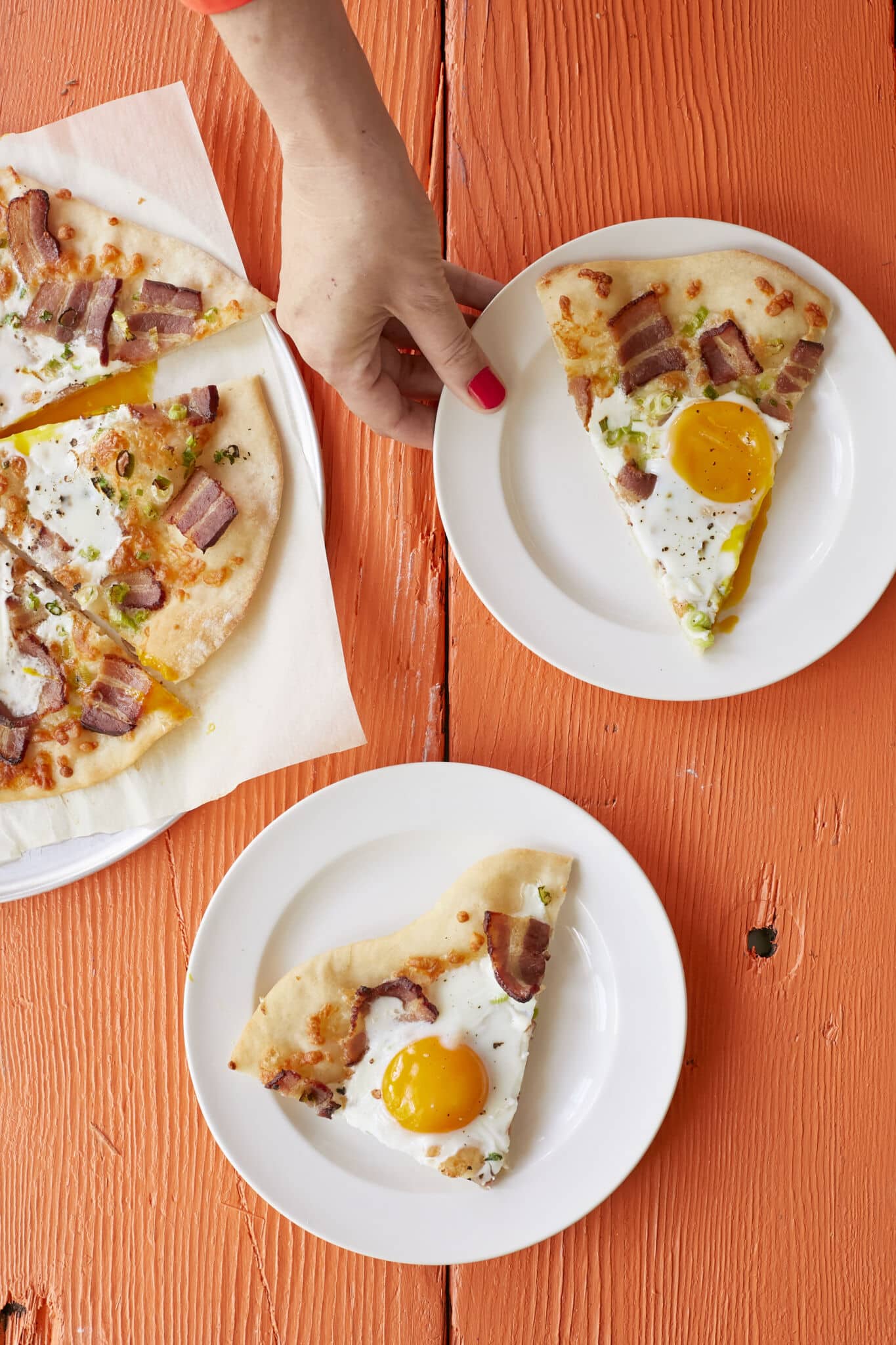 Wholesome breakfast pizza has golden and crispy crust, with smoky bacon, green onions, and sunny-side-up eggs bringing layers of savory flavor. It's sliced and served on two small plates. 