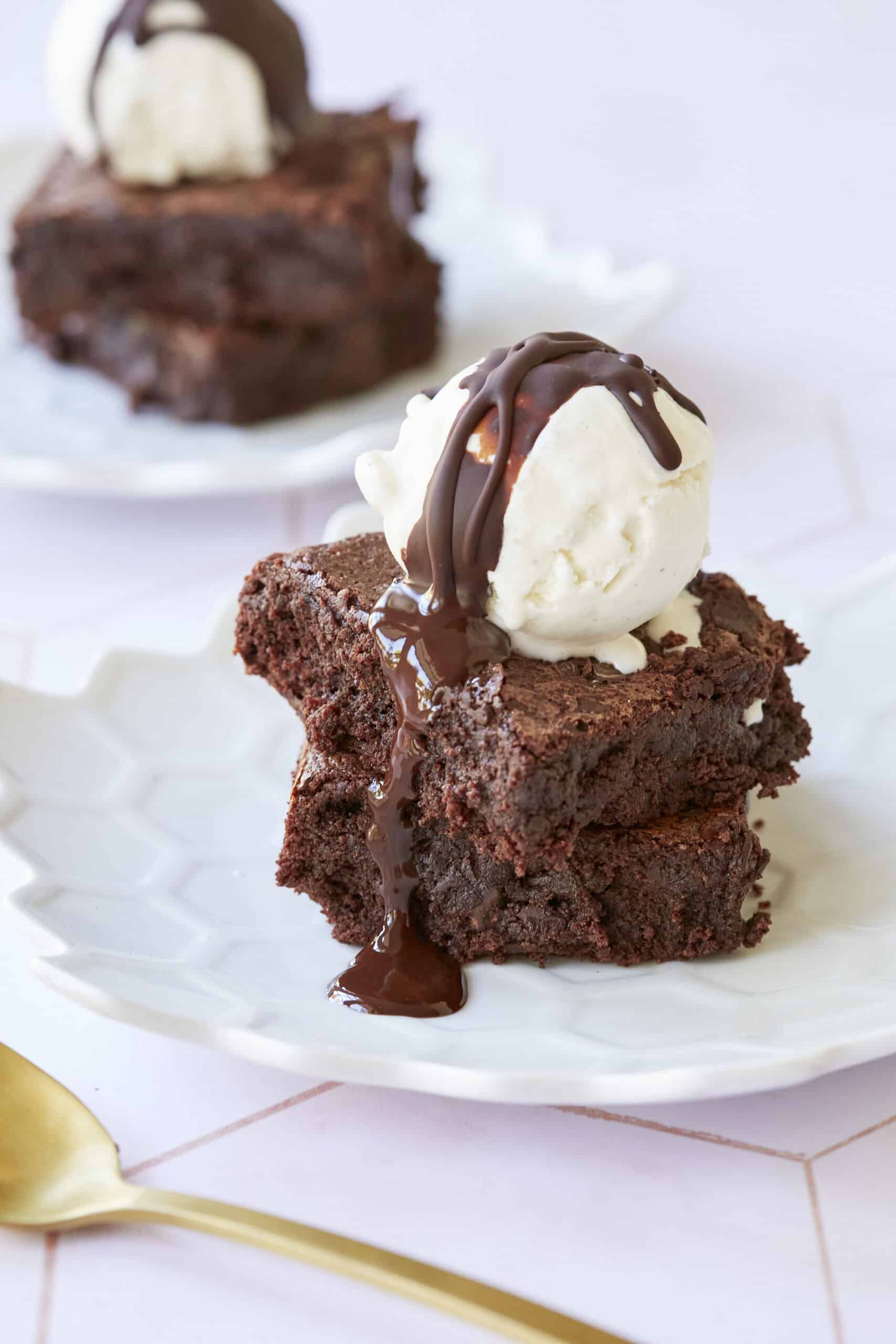 A close-up shot at Gluten-Free Almond Flour Brownies shows the fudge inside and chunks of chocolate with shinny, crinkly top. They're served with ice cream and chocolate sauce. 