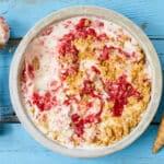 An over-head shot of a bowl of Strawberry Cheesecake Ice Cream shows the creamy, thick ice cream with swirls of jammy strawberries and chunks of buttery Graham cracker crust. An ice cream scoop is on the left side and an ice cream cone is on the right.