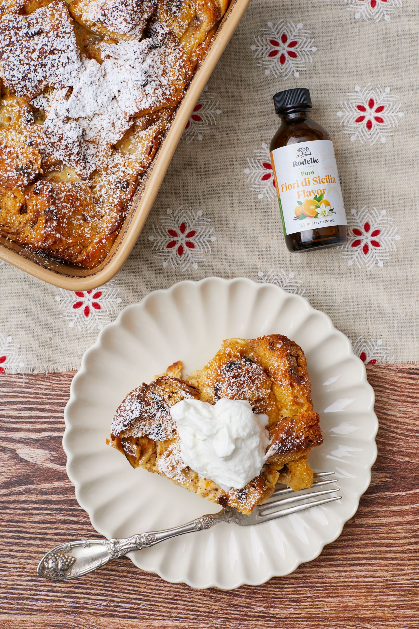 A slice of Panettone Bread Pudding is served with whipped cream on a dessert plate next to the main dish with a bottle of flavor product.