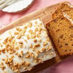 A top down view of a loaf of carrot bread topped with cream cheese glaze and toasted walnuts with two slices have been cut, which shows the moist inside.