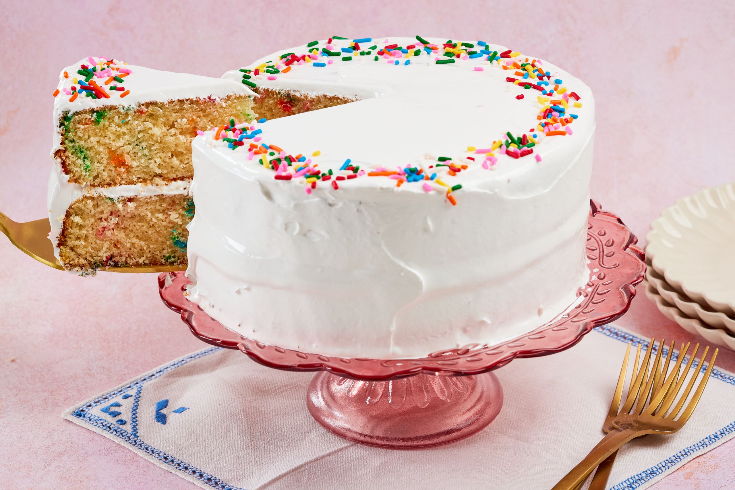 The $5 birthday cake is decorated with white frosting and colorful sprinkles. One slice is cut and being lifted away. It shows the moist crumb and frosting in between.