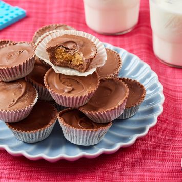 Reese's Peanut Butter cups are staked on a plate, with the top one having one bite taken. It shows the rich filling. Milk is served on the side.