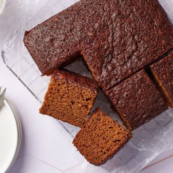 A molasses cake has been cut into 9 big squares on the cooling rack. Two are having the sides facing up, showing the moist and airy crumb. Whipped cream is served on the side.