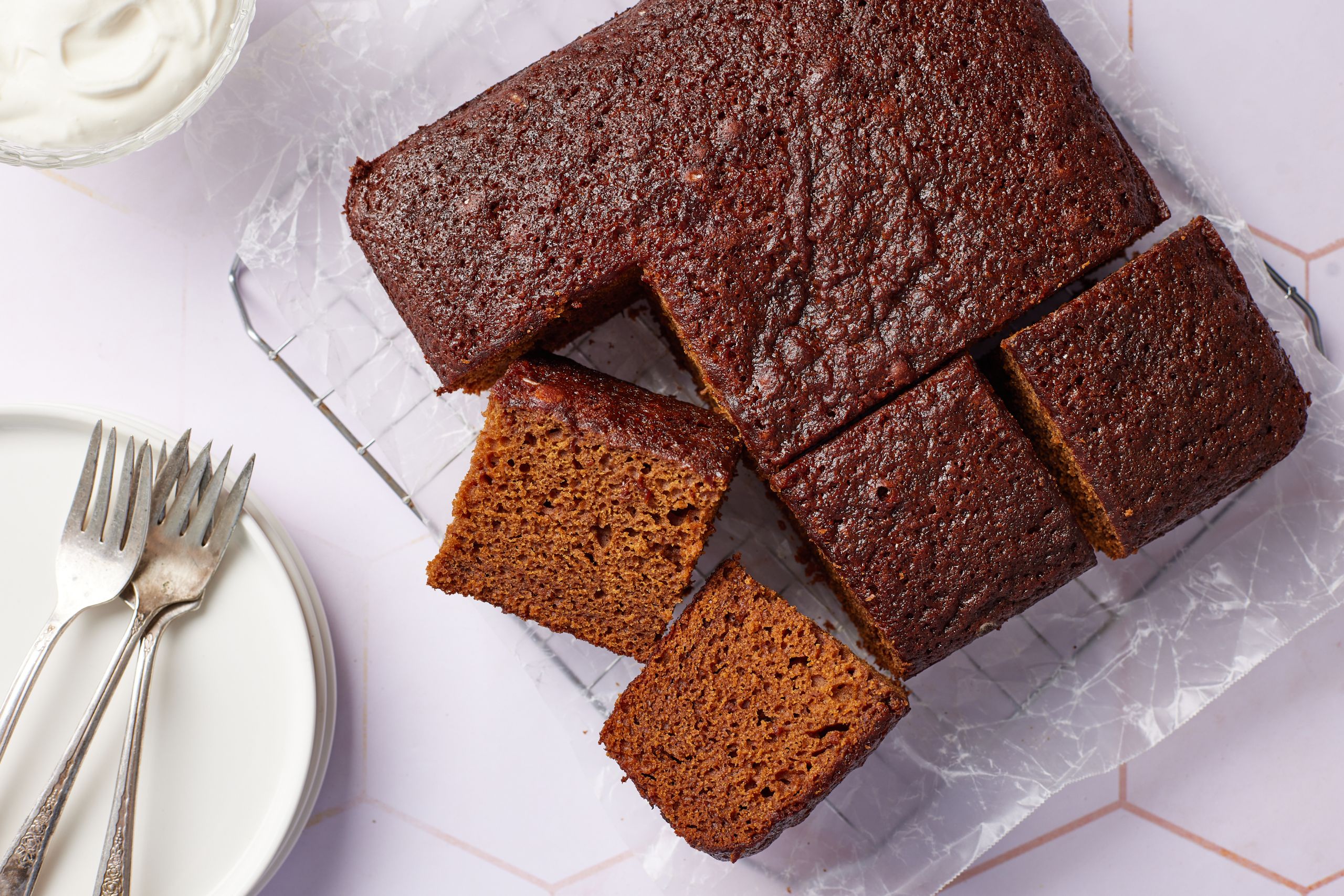 A molasses cake has been cut into 9 big squares on the cooling rack. Two are having the sides facing up, showing the moist and airy crumb. Whipped cream is served on the side.