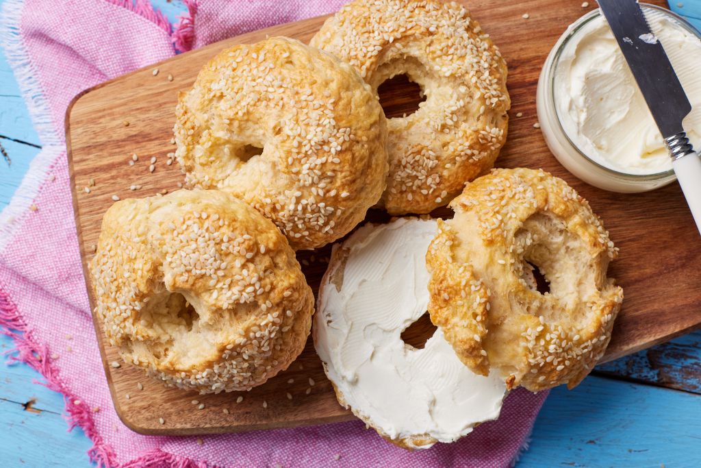 4 golden bagels are on the wooden board. One is sliced in half and served with cream cheese.
