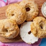 4 golden bagels are on the wooden board. One is sliced in half and served with cream cheese.